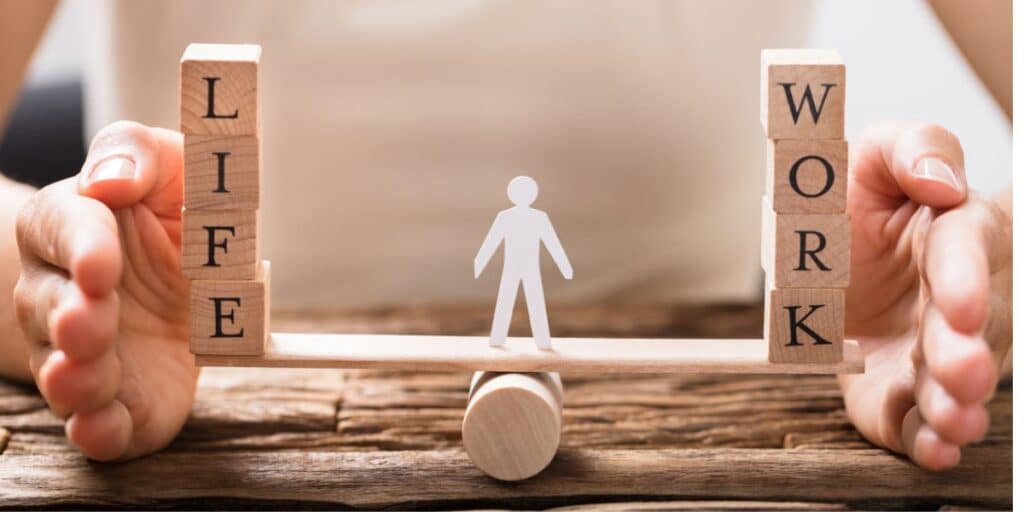 Wooden blocks spelling 'LIFE' and 'WORK' balanced on a seesaw with a small figure in the center, symbolizing the concept of work-life balance