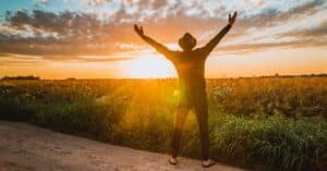 A man raises arms in freedom at sunrise in a rural field, expressing joy and connection with nature.