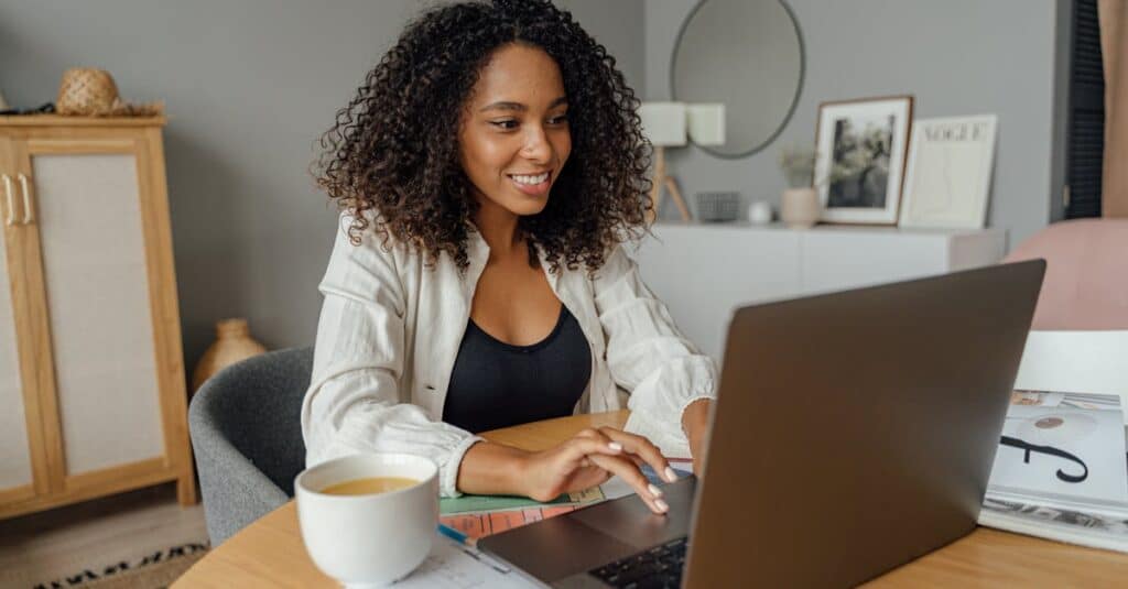 A woman sits at a round table, working on a laptop with a coffee cup nearby in a cozy home office setting.